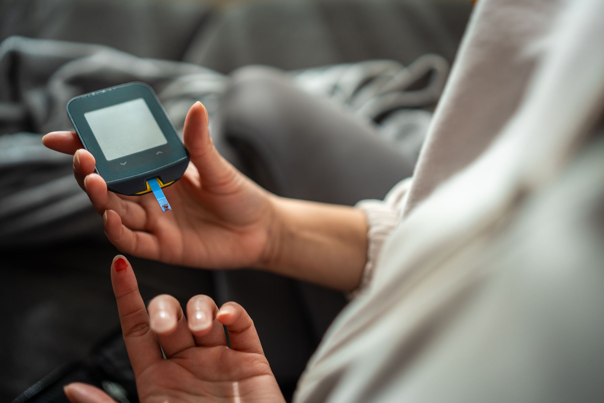 Close-up view of a female's hand using a glucometer to check her blood sugar level. The setting is indoors, with the focus on the device and the blood sample on her fingertip. Her attire is casual, reflecting a home healthcare activity.